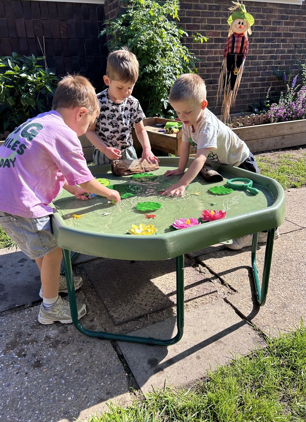 Three children exploring a water tray with toy frogs and lily pads